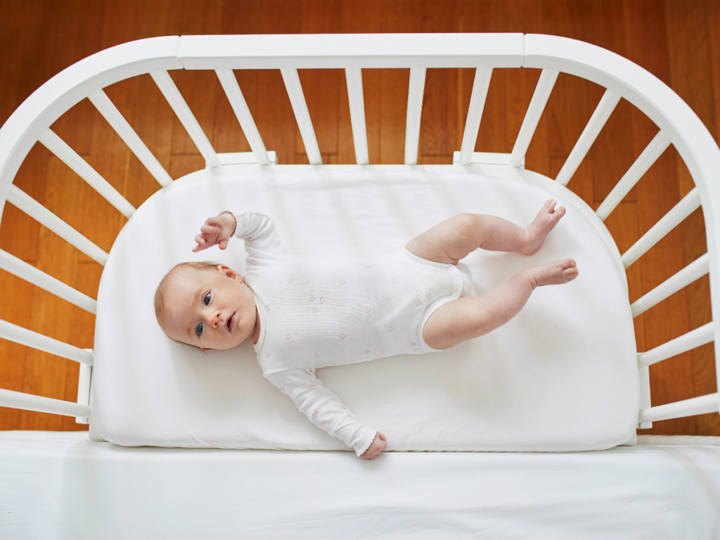 Baby lying on a firm white mattress inside a co sleeper bassinet.